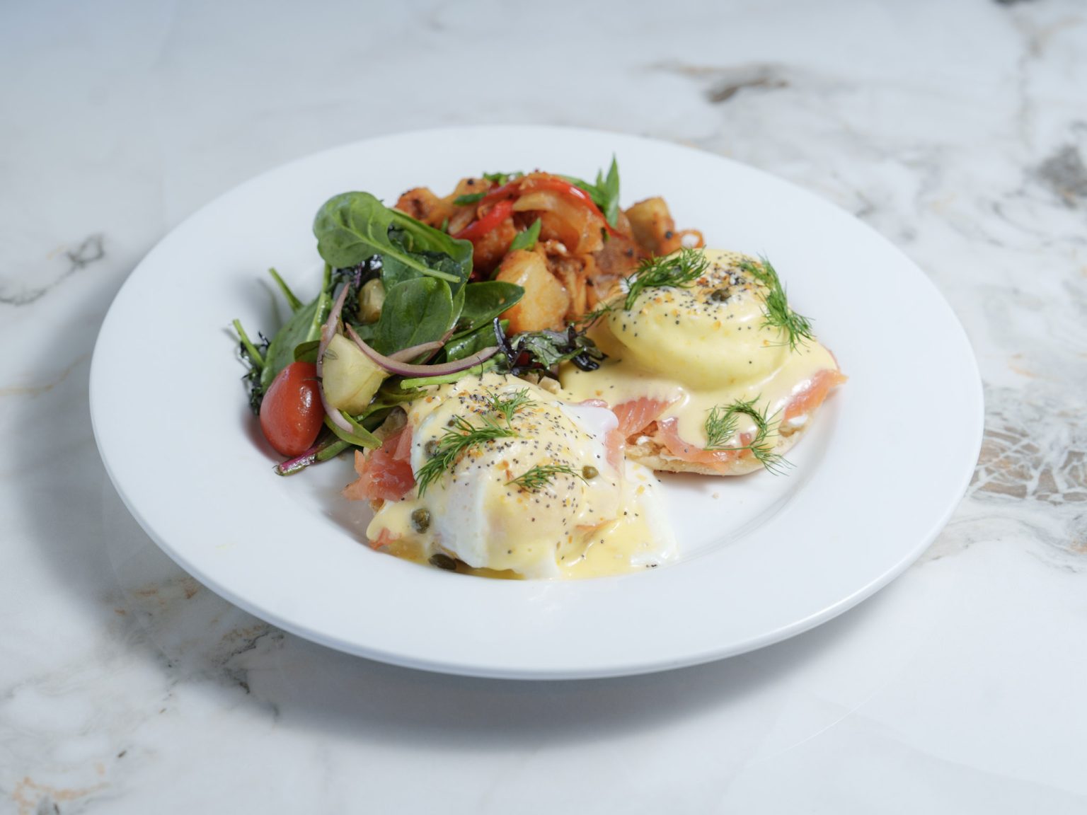 Gourmet smoked salmon Eggs Benedict served with fresh greens and roasted potatoes on a white plate.