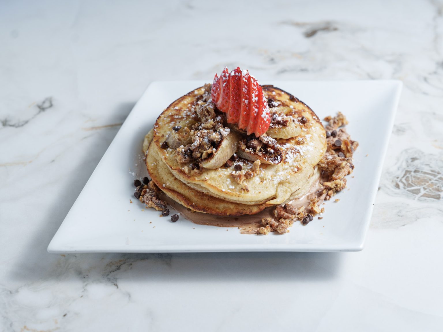Gourmet sweet pancakes served on a white plate with fresh strawberries, sliced bananas, and chocolate crumble.